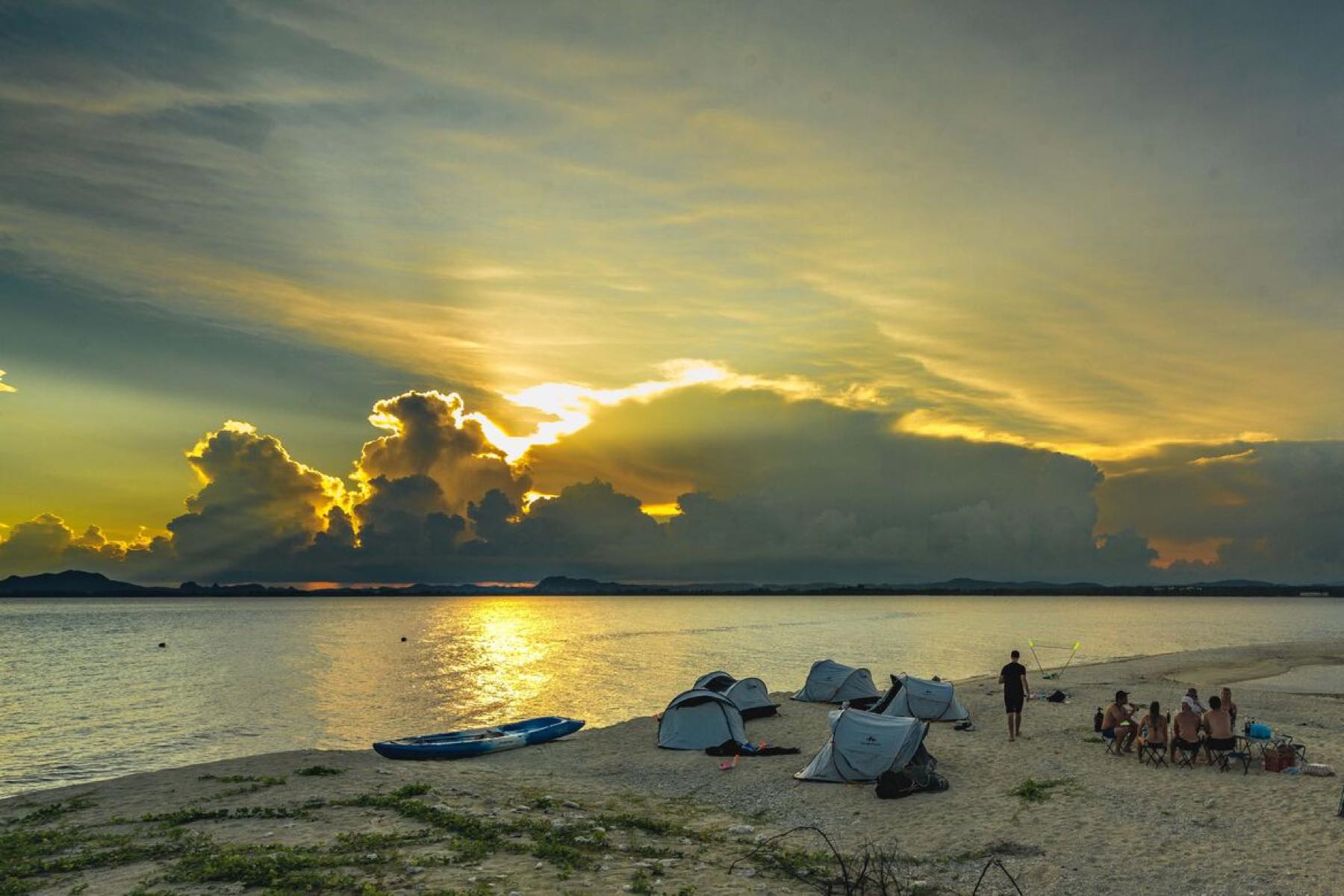 Sunset over the sea with tents pitched on a sand spit and a few small figures on the shore.